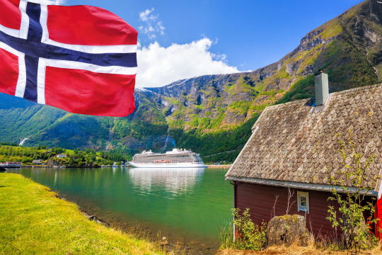 Links eine norwegische Fahne, im Hintergrund im einem Fjord gelegen ein weißes Kreuzfahrtschiff, rechts ein rotes Holzhaus